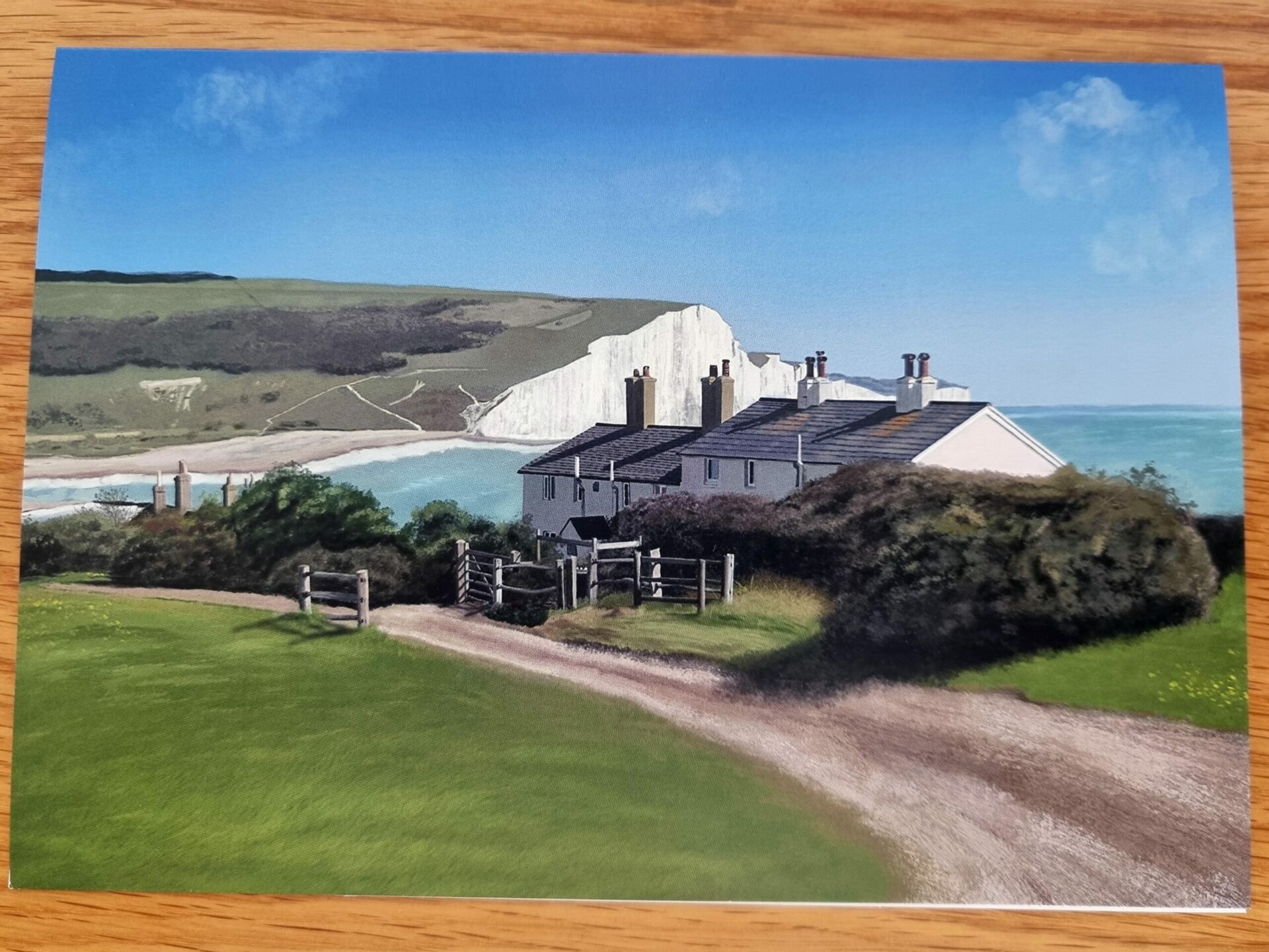 Products In Real Life 7 Coastal landscape featuring charming cottages near the sea, with white chalk cliffs in the background and a clear blue sky. A winding dirt path leads through lush greenery, highlighting the serene beauty of the countryside.
