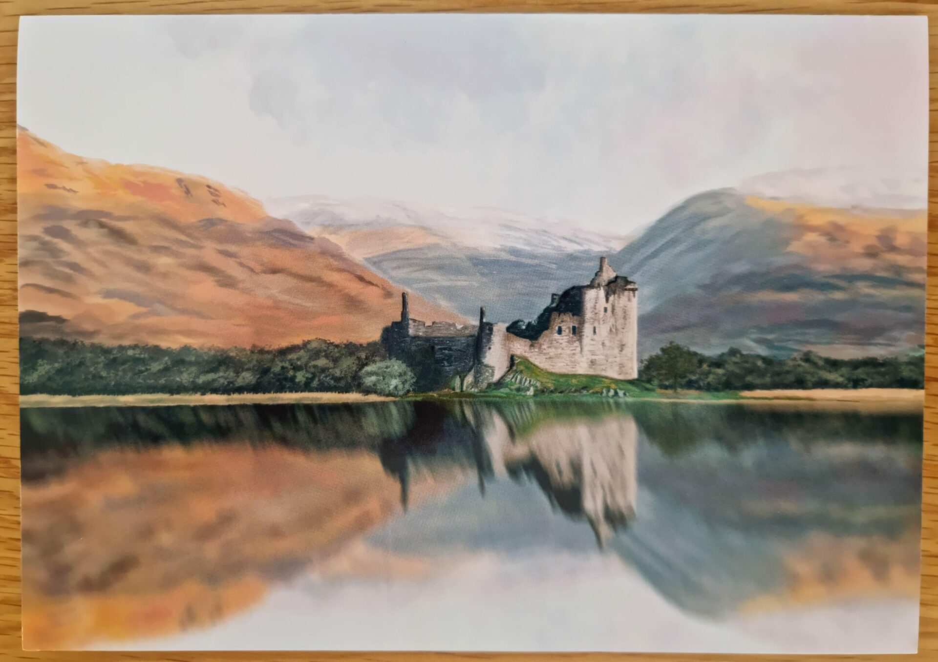 Products In Real Life 9 Scenic view of a historic castle on the edge of a calm lake, surrounded by lush greenery and rolling hills, with mountains in the background reflecting in the water. Ideal for showcasing natural beauty and architectural history.